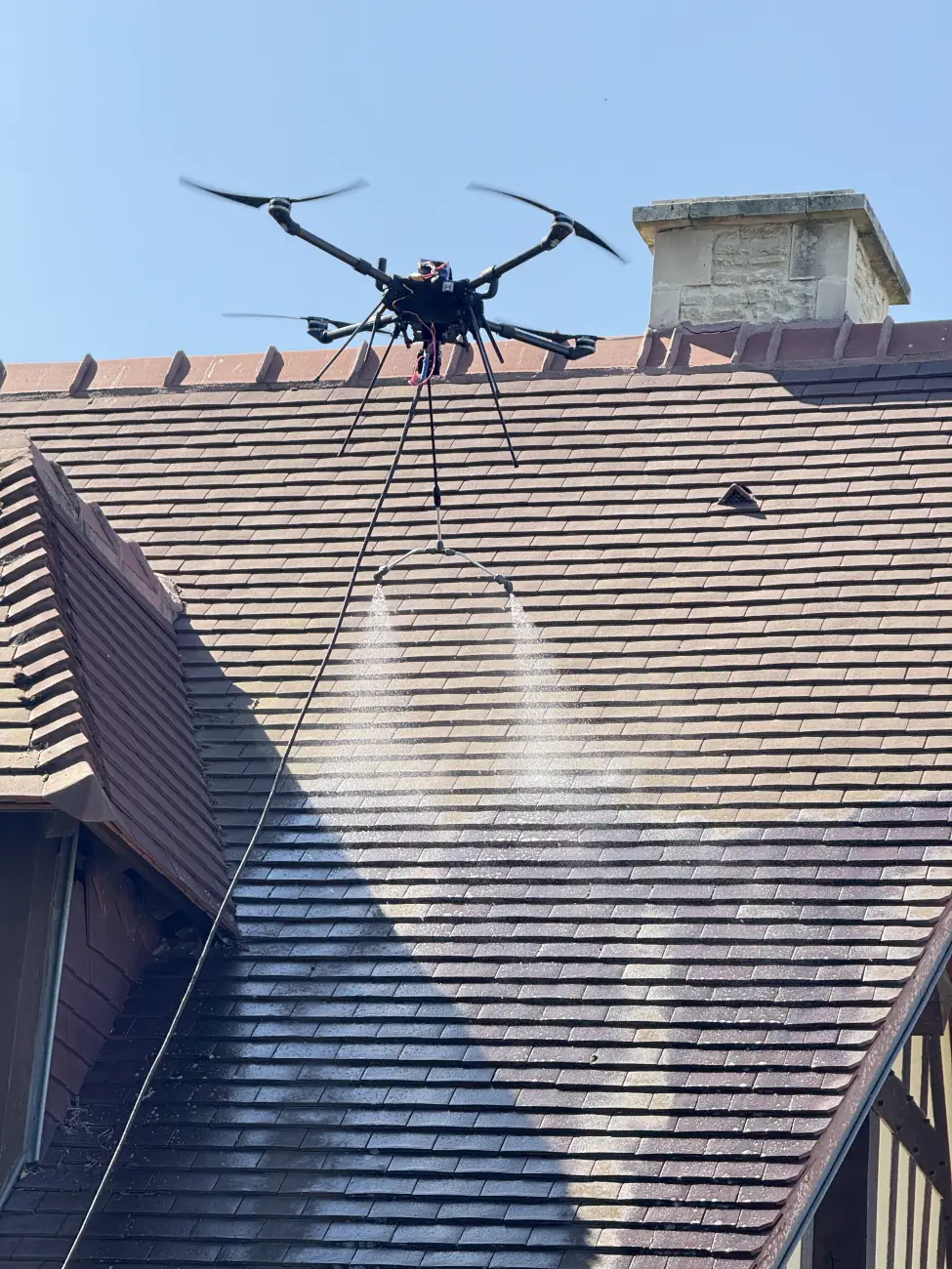 Drone équipé d'une double buse pulvérisant un traitement démoussant sur la toiture en tuiles d'une maison normande à colombages