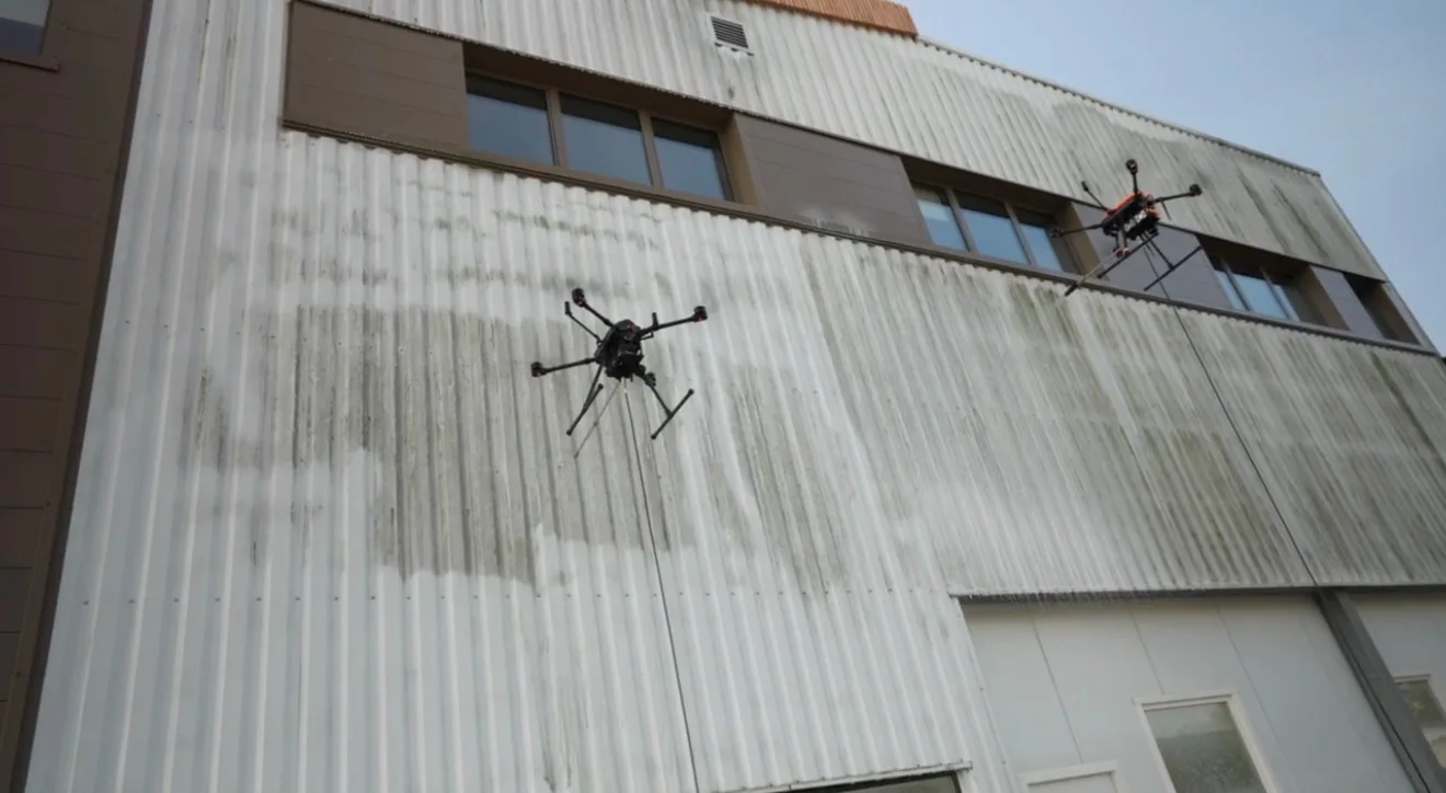 Deux drones de nettoyage travaillant simultanément sur la façade industrielle encrassée d'un grand bâtiment : cadence doublée sur les grandes surfaces