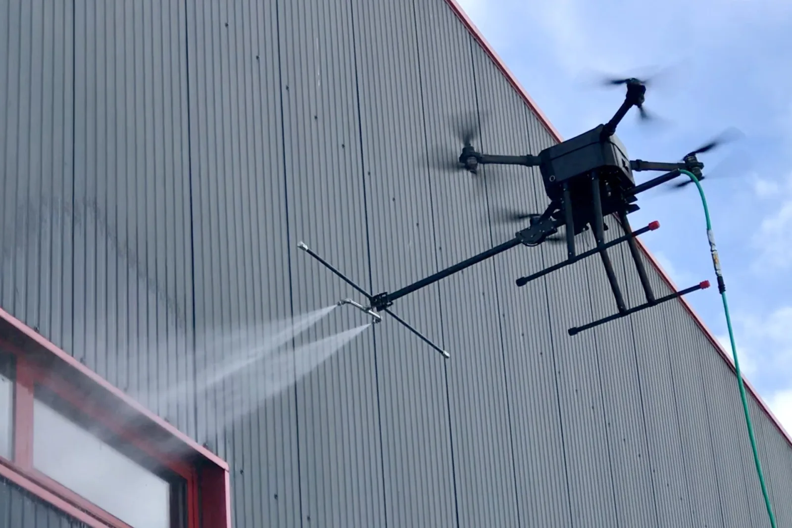 Nettoyage d'un bardage métallique industriel par drone pulvérisateur : jet appliqué au plus près de la paroi du hangar, sans nacelle ni immobilisation de l'activité